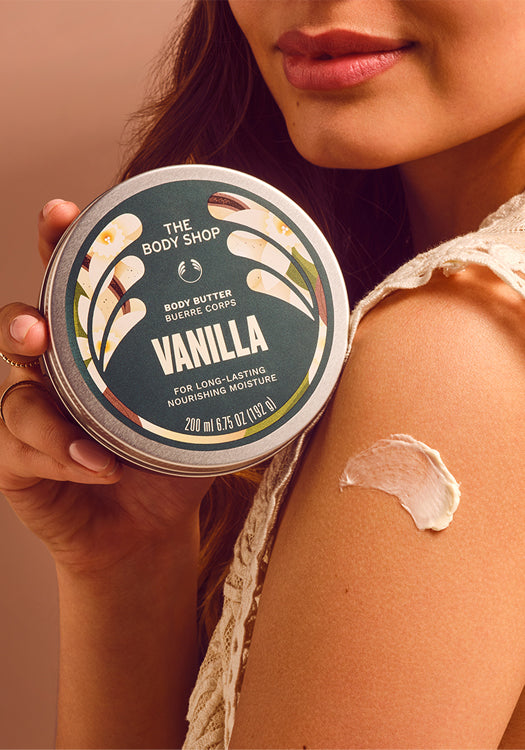 A close-up lifestyle shot of a woman holding a tin of The Body Shop Vanilla Body Butter, with a dollop of the cream-colored body butter visible on her shoulder against a warm beige background.