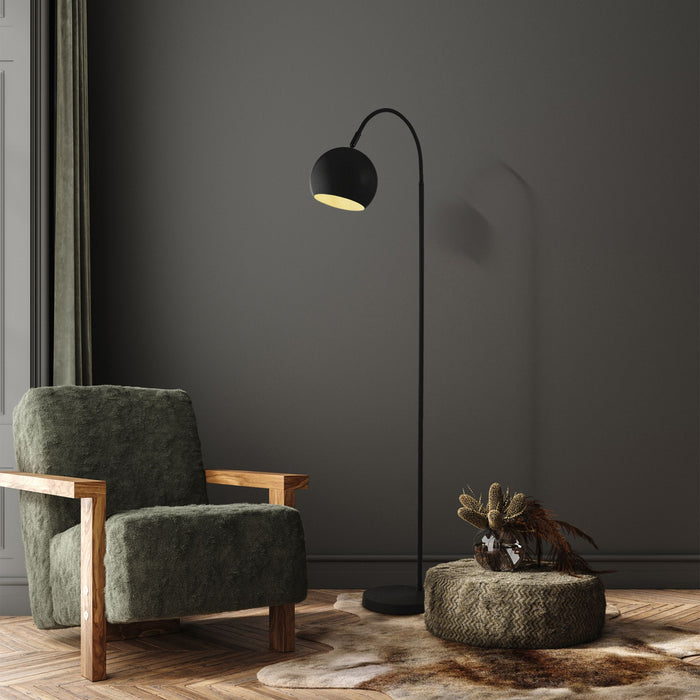 Calumn black arc floor lamp lit in a dark grey living room beside a green armchair and woven pouffe
