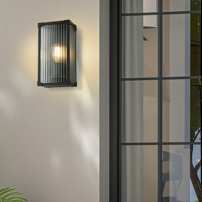 Cincinnati black ribbed glass wall lantern lit on a white wall beside a dark framed window