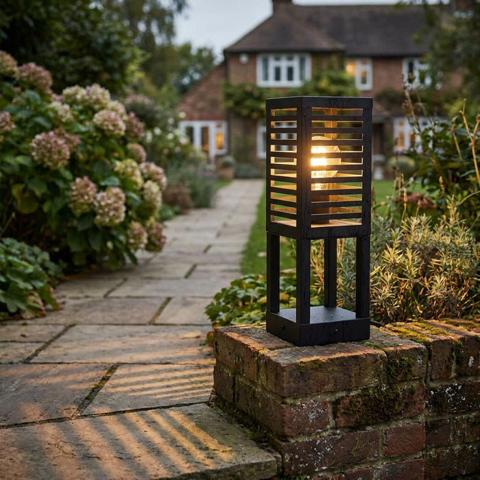 Modern black slatted garden pedestal light glowing on a brick wall, casting shadows across a stone garden path.