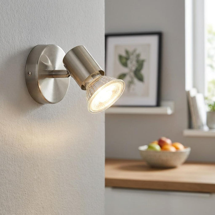 Eglo Buzz-LED satin nickel wall spotlight mounted on a textured wall, illuminating a blurred kitchen setting.