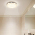 A round, white LED flush ceiling light illuminating a modern bathroom with beige wall tiles.