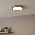 Round nickel flush mount LED ceiling light illuminating a neutral-toned room above a wooden shelf.