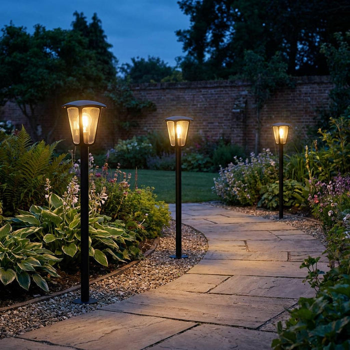 Three black bollard lights illuminating a winding stone garden path at twilight among lush green plants.