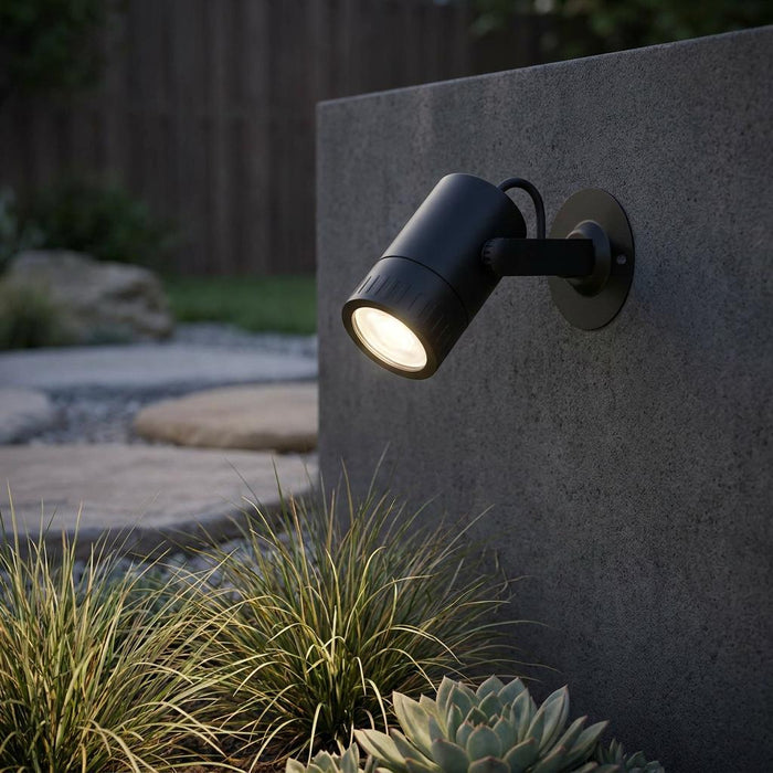 Black outdoor smart LED spotlight mounted on a gray concrete wall, illuminating garden plants at dusk.