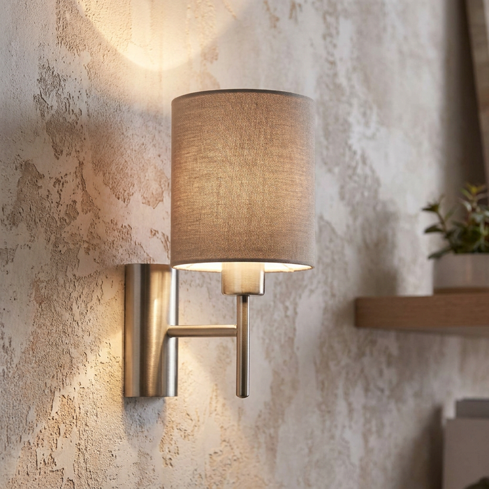 Brushed nickel wall sconce with a taupe cylindrical fabric shade mounted on a rustic textured wall.