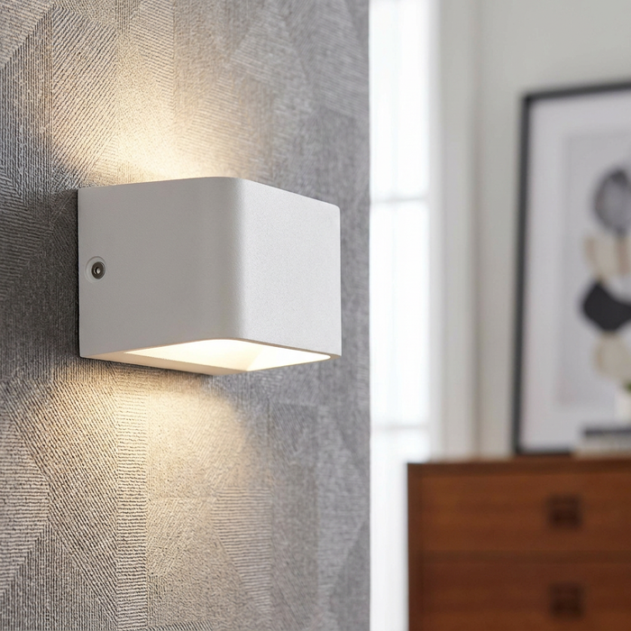Modern white cube wall light casting a warm up-and-down glow on a grey textured geometric wallpaper.