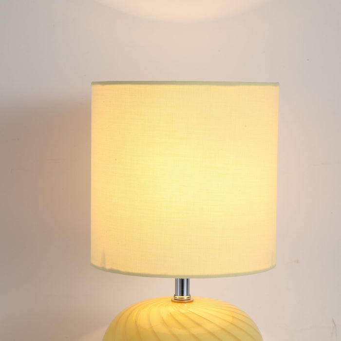Close-up of the Fete lamp glowing cream shade above its swirled yellow glass base