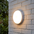 Circular white LED bulkhead light glowing on an exterior white brick wall.
