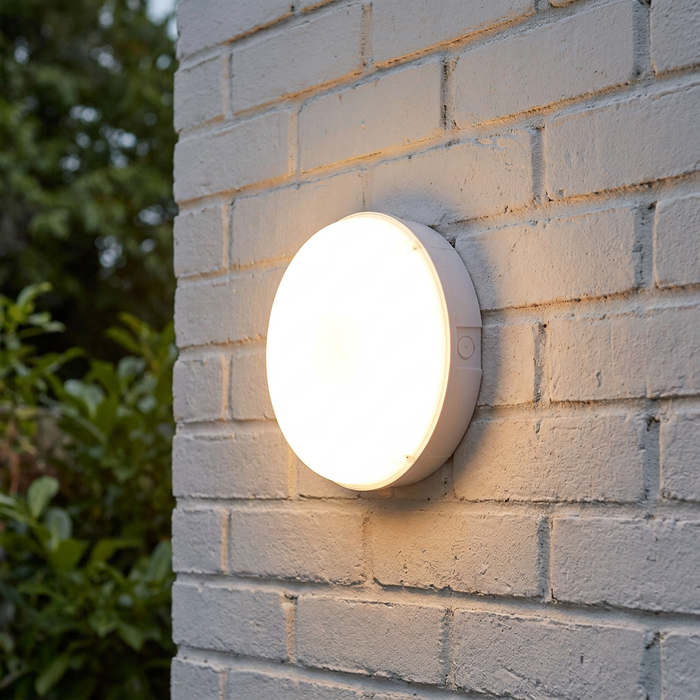 Circular white LED bulkhead light glowing on an exterior white brick wall.