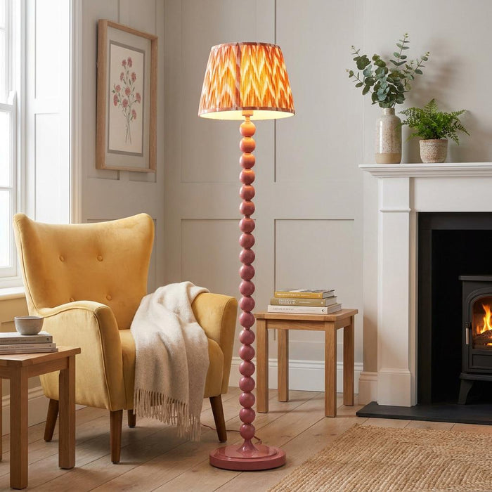 Pink bobbin floor lamp with an orange zigzag shade in a cozy living room next to a yellow armchair and fireplace.