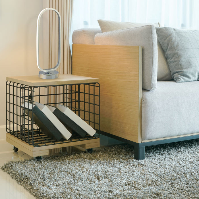Liner chrome LED loop lamp lit on an oak side table beside a grey sofa and wire magazine rack