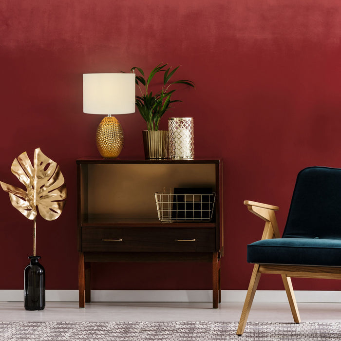 Nadine gold lamp on a walnut cabinet against a deep red wall with teal armchair and plants