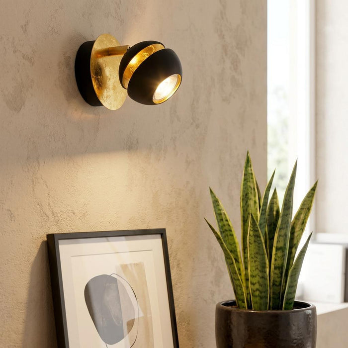 Modern black and gold leaf wall spotlight illuminating a textured interior wall with a plant and picture frame.