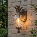 Pineapple-shaped bronze outdoor wall lantern with textured glass glowing on a light brick wall.