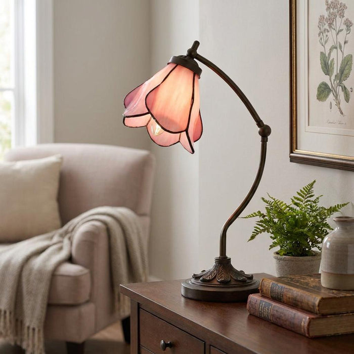 A pink tulip-style Tiffany desk lamp with a bronze swan neck stand lit on a wooden table in a living room.