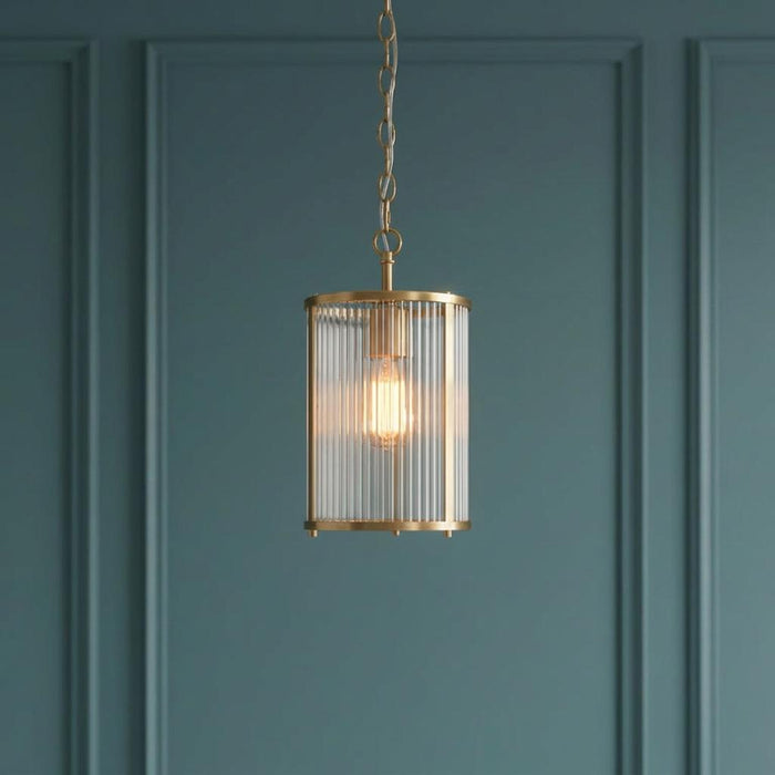 An illuminated antique brass pendant light with a cylindrical ribbed glass shade against a dark teal paneled wall.