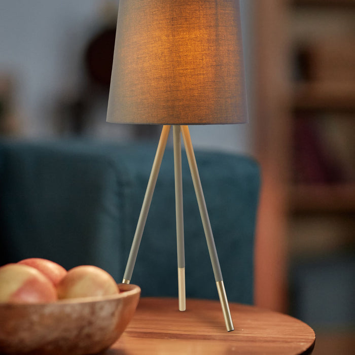 Rio tripod lamp lit on a wooden side table beside a fruit bowl and teal sofa