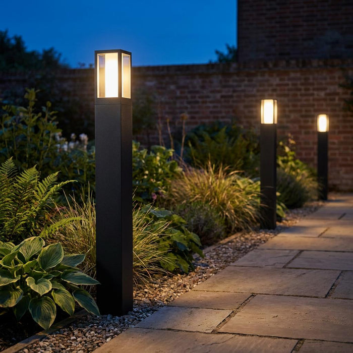 Three black square bollard lights illuminating a stone garden path at twilight with a warm glow.
