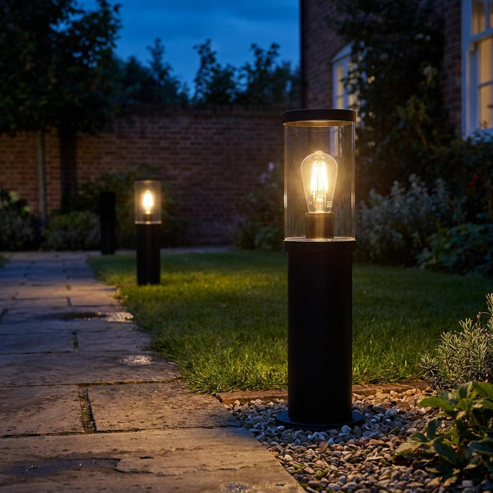 Two black outdoor bollard lights with warm bulbs lighting up a garden path and lawn at twilight.