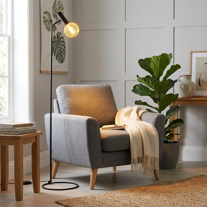 Black floor lamp with a mesh globe bulb illuminating a grey armchair in a cozy living room corner.