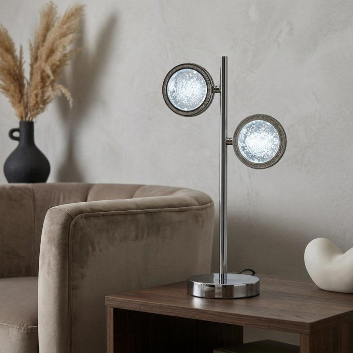 Modern chrome table lamp with two circular quartz-effect glass shades on a wooden side table in a living room.