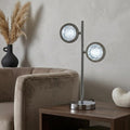 Modern chrome table lamp with two circular quartz-effect glass shades on a wooden side table in a living room.