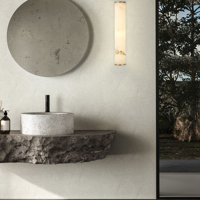 Concrete bathroom with the Wales gold wall light beside a round mirror and stone basin